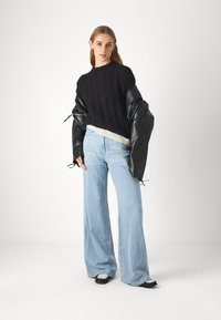 Maglione nero a trama cable-knit con orlo grigio chiaro, indossato con jeans azzurri a gamba larga e maniche oversize in pelle nera. Scarpe nere con accenti bianchi.