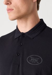 Polo preto feito de algodão, com um colarinho texturizado, cinco botões e um logótipo cinza no peito com um design de corda.