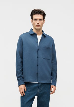 Junger Mann, der ein blaues Hemd mit Brusttasche über einem weißen T-Shirt und blauen Jeans trägt, steht vor einem schlichten hellen Hintergrund.