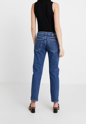 Kobieta ubrana w niebieskie dżinsy z wysokim stanem i czarny bezrękawnik, stojąca tyłem na jednolitym białym tle.