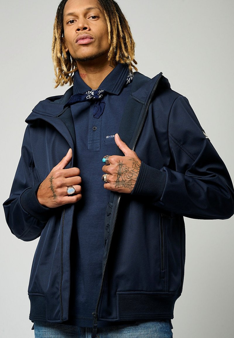 Veste bleu marine avec fermeture éclair à l'avant, capuche et poignets côtelés. Le tissu semble lisse, porté sur un polo bleu foncé. Tatouages remarquables sur les mains.