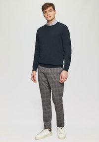 Marineblauwe crew-neck trui over een lichtblauw overhemd met kraag, gecombineerd met grijs geblokte broek en witte sneakers met grijze accenten.