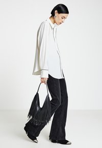 Donna che indossa una camicetta bianca con rifiniture nere, jeans neri a zampa e stivali neri con frange, portando una borsa nera con frange.