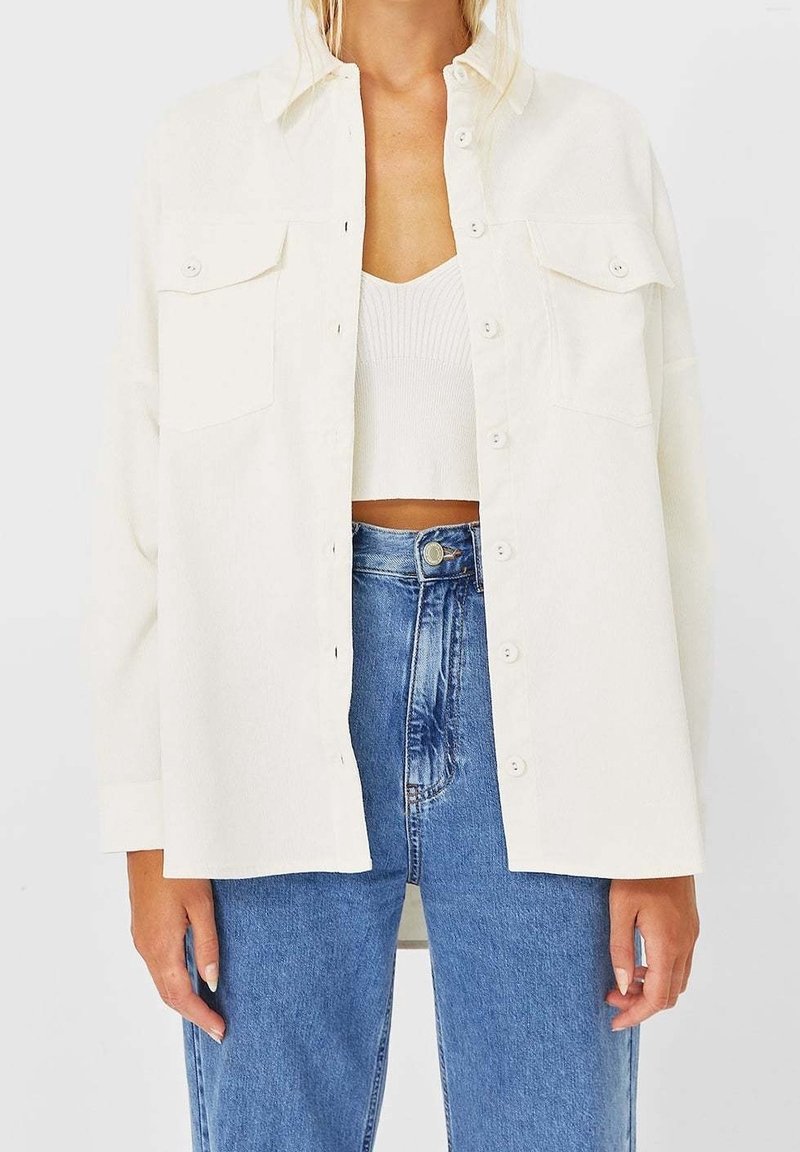 White button-up shirt with two chest pockets, soft texture, and a relaxed fit, paired with high-waisted blue denim jeans.