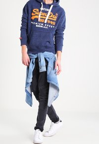 Sudadera de marina con logotipo naranja, pantalones jogger negros, camisa de denim atada a la cintura y zapatillas blancas. Diseño sencillo con texturas contrastantes.