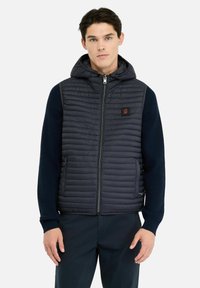 Gilet imbottito blu navy con cappuccio, caratterizzato da una texture trapuntata, chiusura con zip e tasche laterali. Le maniche in maglia a coste aggiungono contrasto.