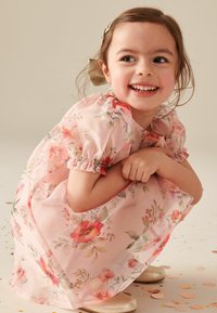 Leende ung flicka som sitter på huk, iklädd en rosa blommig klänning, guldskor och en guldkam, med konfetti spritt på golvet.