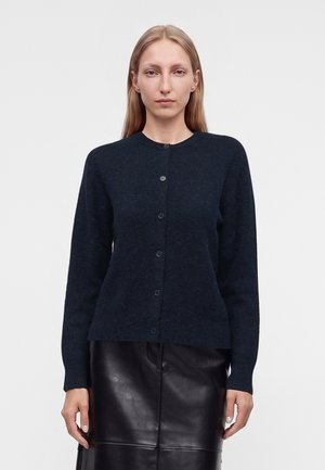 Vrouw met recht blond haar draagt een donkerblauwe gebuttonde trui en een zwarte leren rok, staand tegen een effen witte achtergrond.