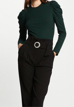 Vrouw die een donkergroene geribde lange mouwen top draagt met opgepofte schouders en een zwarte hooggetailleerde broek met een decoratieve riem met ronde gesp.