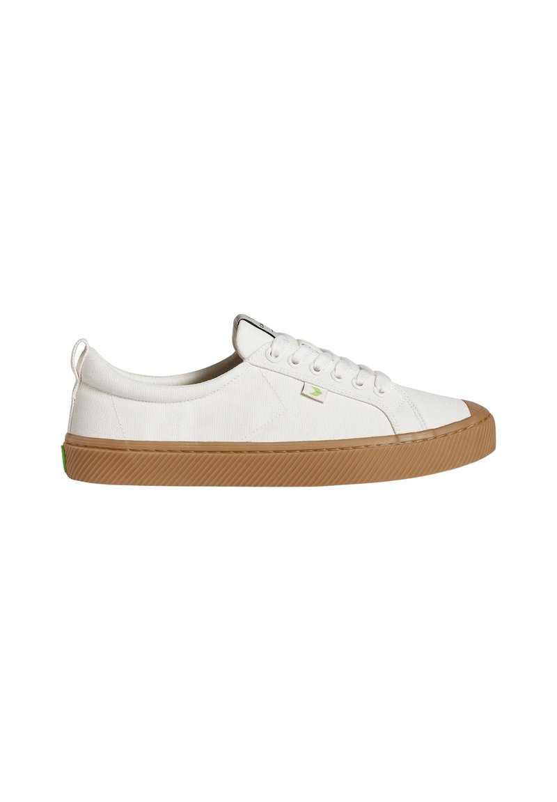 Sneakers bianche in tela con una suola in gomma marrone testurizzata, dotate di sei asole per i lacci, un logo verde di accentuazione e un piccolo tab sul tallone.