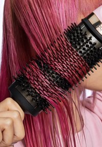 Okrągła czarna szczotka do włosów z miękkim włosiem, trzymana przy długich różowych włosach. Szczotka ma teksturowany uchwyt i ergonomiczny chwyt.