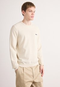 Giovane uomo con capelli chiari che indossa un maglione a girocollo color crema con il logo "BOSS" e pantaloni beige a pieghe, in piedi davanti a uno sfondo bianco semplice.