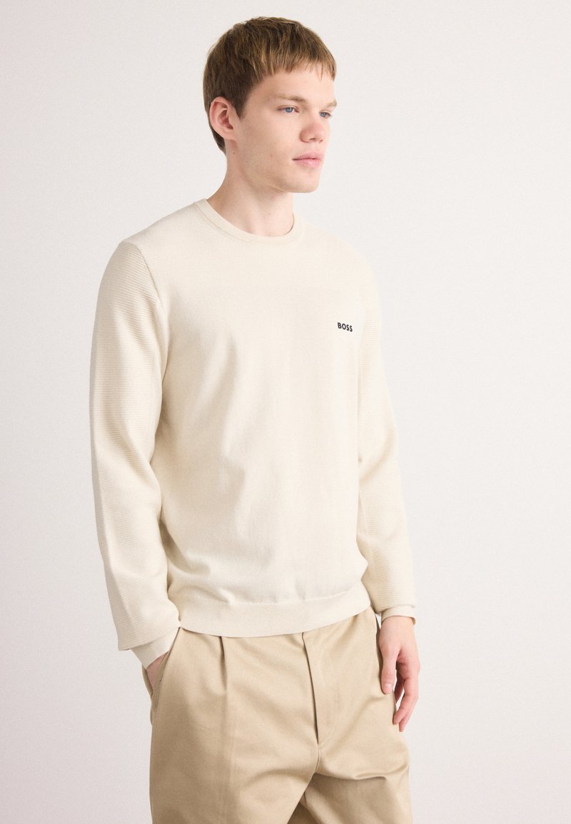 Giovane uomo con capelli chiari che indossa un maglione a girocollo color crema con il logo "BOSS" e pantaloni beige a pieghe, in piedi davanti a uno sfondo bianco semplice.