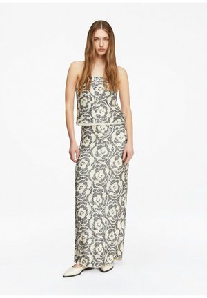 Frau mit langen, glatten Haaren, die ein ärmelloses Oberteil und einen langen Rock mit schwarz-weißem Blumenmuster trägt, steht mit entspannten Händen.