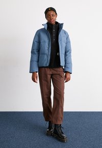 Blå puffjakke med høj krave, parret med en sort fleecepullover, brune straight-leg bukser og sorte snøresko.