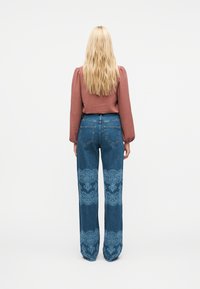 Γυναίκα με μακριά ξανθά μαλλιά, φορεμένη με μωβ μπλούζα μακρυμάνικη και μπλε τζιν με δαντελωτά σχέδια στο κάτω μέρος των ποδιών, που στέκεται γυρισμένη προς τα πίσω.