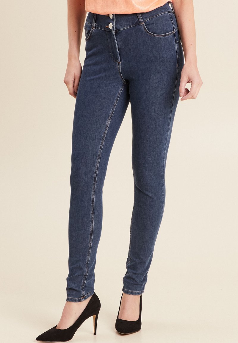 Jean skinny bleu foncé en denim avec une texture lisse, présentant une taille mi-haute et des détails de couture subtils. Associé à des talons.