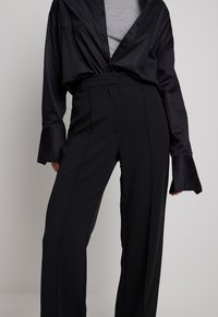 Schwarze Satinbluse mit lockerem Schnitt und Knopfleiste, kombiniert mit hochgeschnittenen schwarzen Hosen, die eine glatte Textur und Seitentaschen haben.