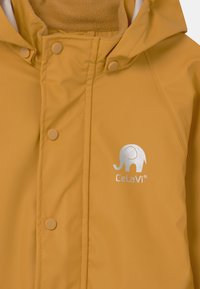 Gul vattentät jacka med texturerad yta, med en silverfärgad elefantlogotyp och tryckknappar. Insidan visar ett mjukt material.