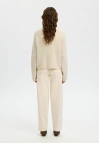Femme aux longs cheveux ondulés portant un pull beige crème en tissu pelucheux, un pantalon beige clair à jambes larges et des chaussures marron, debout devant un fond blanc uni.