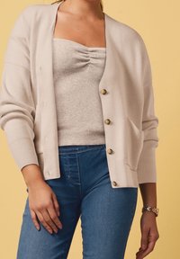 Jasno beżowy, dziany kardigan o luźnym kroju, z złotymi guzikami, ribbowymi mankietami i kieszenią na przodzie, na tle o fakturowanym topie i niebieskich dżinsach.