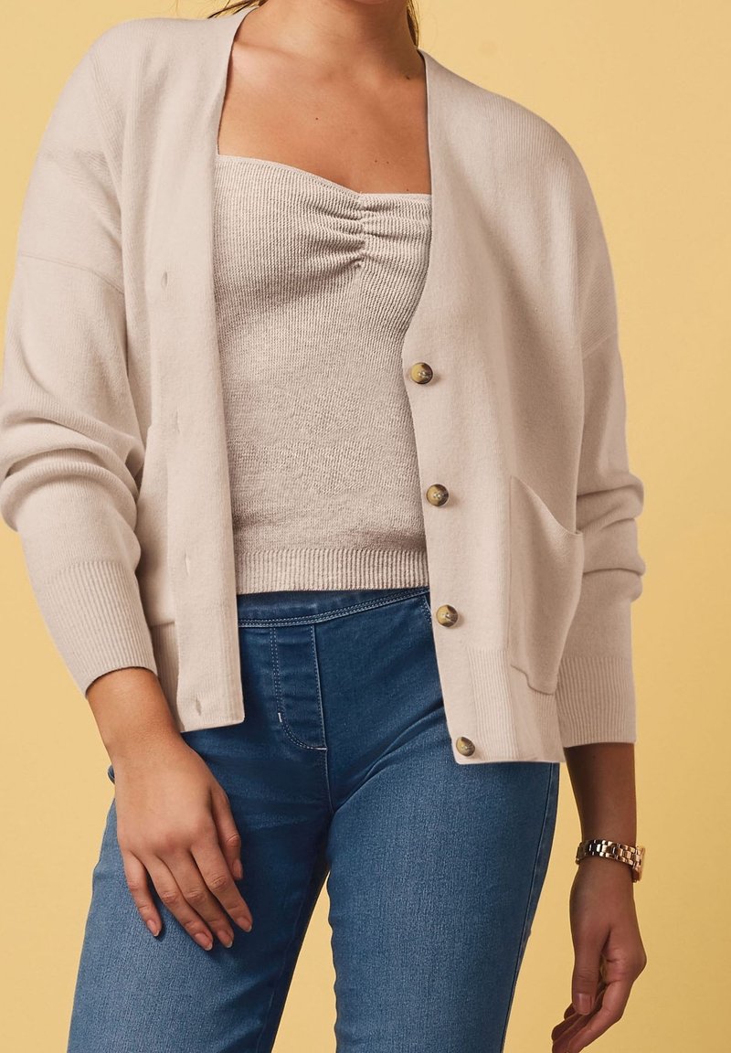 Jasno beżowy, dziany kardigan o luźnym kroju, z złotymi guzikami, ribbowymi mankietami i kieszenią na przodzie, na tle o fakturowanym topie i niebieskich dżinsach.