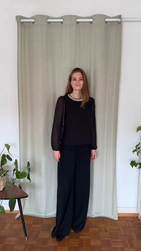 Zwarte blouse met lange mouwen en een halslijn met parels, gecombineerd met zwarte, hoge taille wijde broekspijpen en zwarte schoenen. Effen lichte achtergrond.