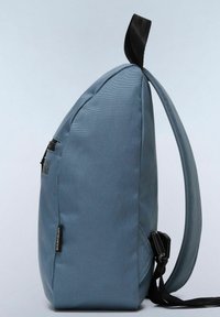 Sac à dos en tissu bleu de forme triangulaire, avec une surface texturée, des accents de fermeture éclair noirs et des bretelles ajustables. La poche latérale est dotée d'un clip.