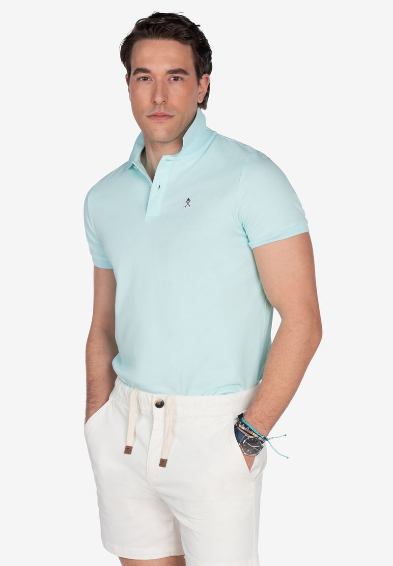 Polo de color azul claro con una tapeta de dos botones y un logo bordado, combinado con pantalones cortos blancos y una pulsera multicolor.