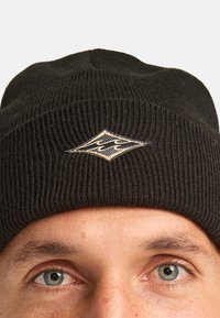 Beanie a coste nera con un patch a forma di diamante caratterizzato da lettering stilizzato in oro e blu navy. Texture morbida.