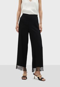 Svarta vida byxor med fransdetalj i nederkant, gjorda av ett mjukt tyg. Kombineras med en vit t-shirt med v-ringning och svarta sandaler med guldiga accenter.