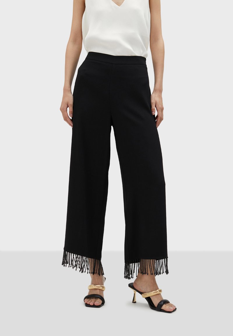 Svarta vida byxor med fransdetalj i nederkant, gjorda av ett mjukt tyg. Kombineras med en vit t-shirt med v-ringning och svarta sandaler med guldiga accenter.