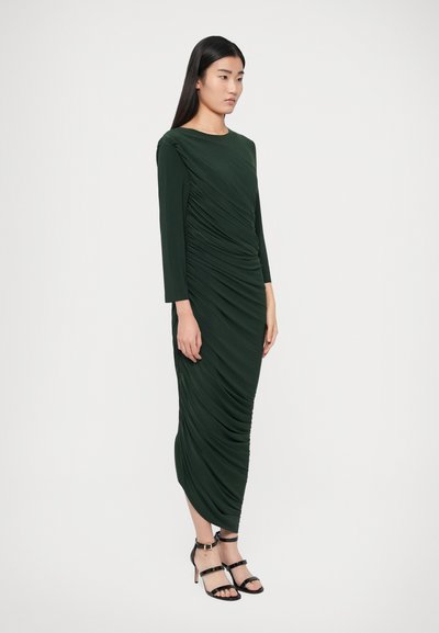 Sukienka w kolorze leśnej zieleni z długim rękawem, marszczonymi detalami, asymetrycznym dołem oraz bocznym drapowaniem. Do tego czarne, wiązane na paski wysokie obcasy.