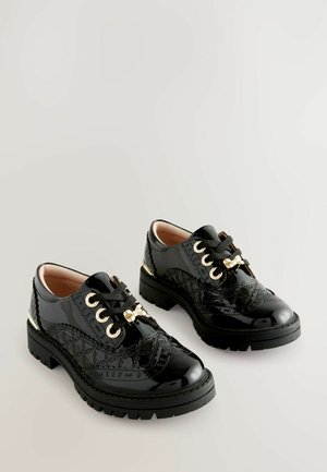 Sorte laklæder sko med en blank finish, præget af guldsmykker, strukturerede detaljer og sorte snørebånd. Kraftig gummisål.