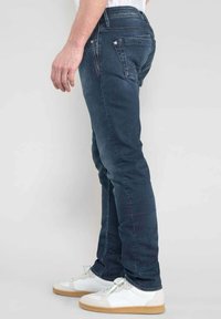 Jeans en denim bleu foncé avec une coupe slim, dotés de cinq poches, de coutures contrastées et d'une taille mi-haute. Portés avec des baskets blanches.