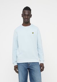 Sweat-shirt bleu clair en coton avec poignets et ourlet côtelés, arborant un petit logo jaune en forme de papillon sur la poitrine gauche. Porté avec un jean bleu.