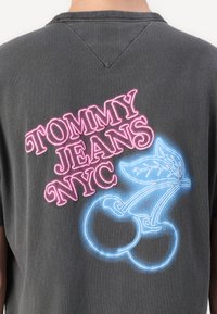 Pilka medvilninė marškinėlė su užrašu "TOMMY JEANS NYC" rožine spalva ir mėlynu neoniniu vyšnios grafiniu motyvu ant nugaros. Trumpomis rankovėmis ir laisvu siluetu.