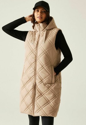 Kobieta w beżowej pikowanej kamizelce z kapturem na czarnym długim rękawie, z jedną ręką w kieszeni i czarną czapką.