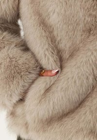 Beige fuskpälsjacka med en mjuk, fluffig textur. Närbild av framsidan, där en hand är insvept i tyget och visar en guldring.