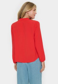 Blusa rossa a maniche lunghe con colletto arricciato. Tessuto morbido, vestibilità comoda e orlo arrotondato. Abbinata a pantaloni blu a gamba larga.