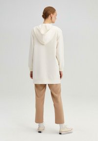 Hanorac de culoare crem, cu o cusătură centrală pe spate și glugă ajustabilă. Purtați cu pantaloni bej și pantofi albi. Textură netedă.