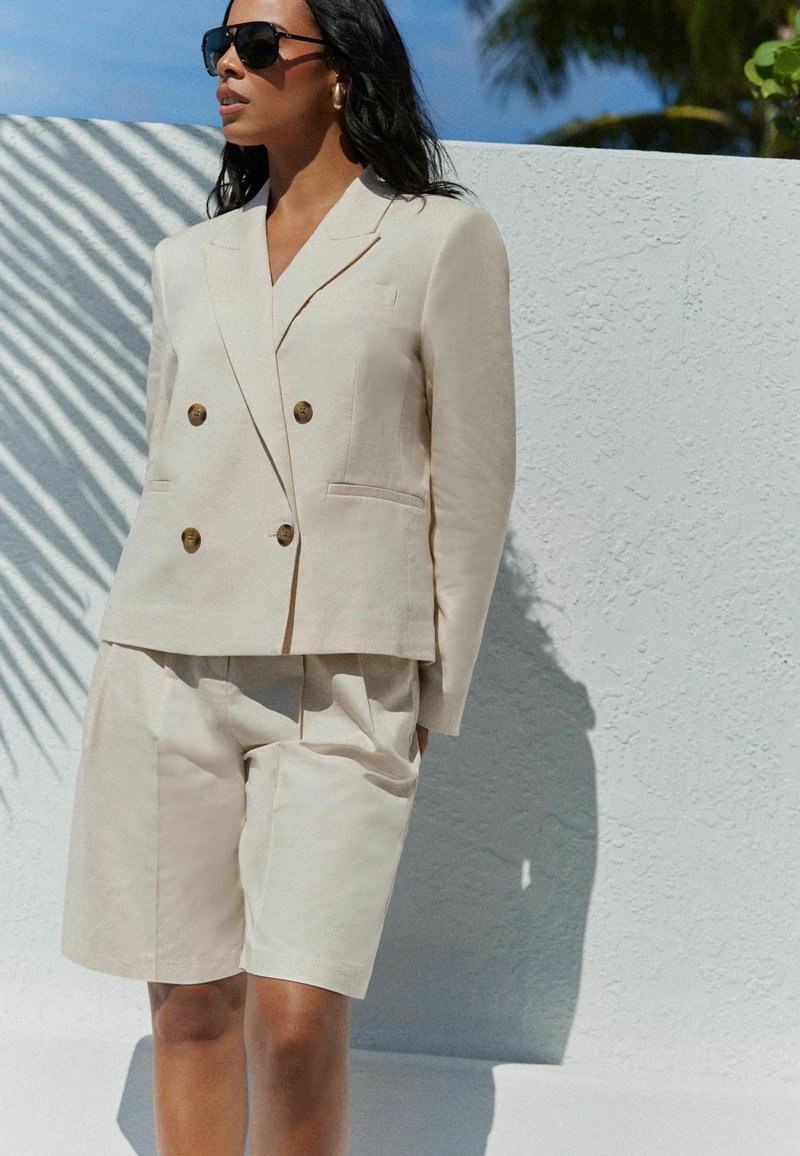 Beige dobbeltradet blazer med messingknapper, parret med matchende skræddersyede shorts; glat tekstur, lommer, sat mod en hvid væg.