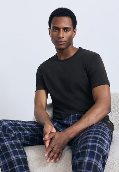 Černé tričko s krátkým rukávem spojené s modrými károvanými pyžamovými kalhotami. Model sedí a předvádí texturu látky a vzory. Jednoduché designové prvky.