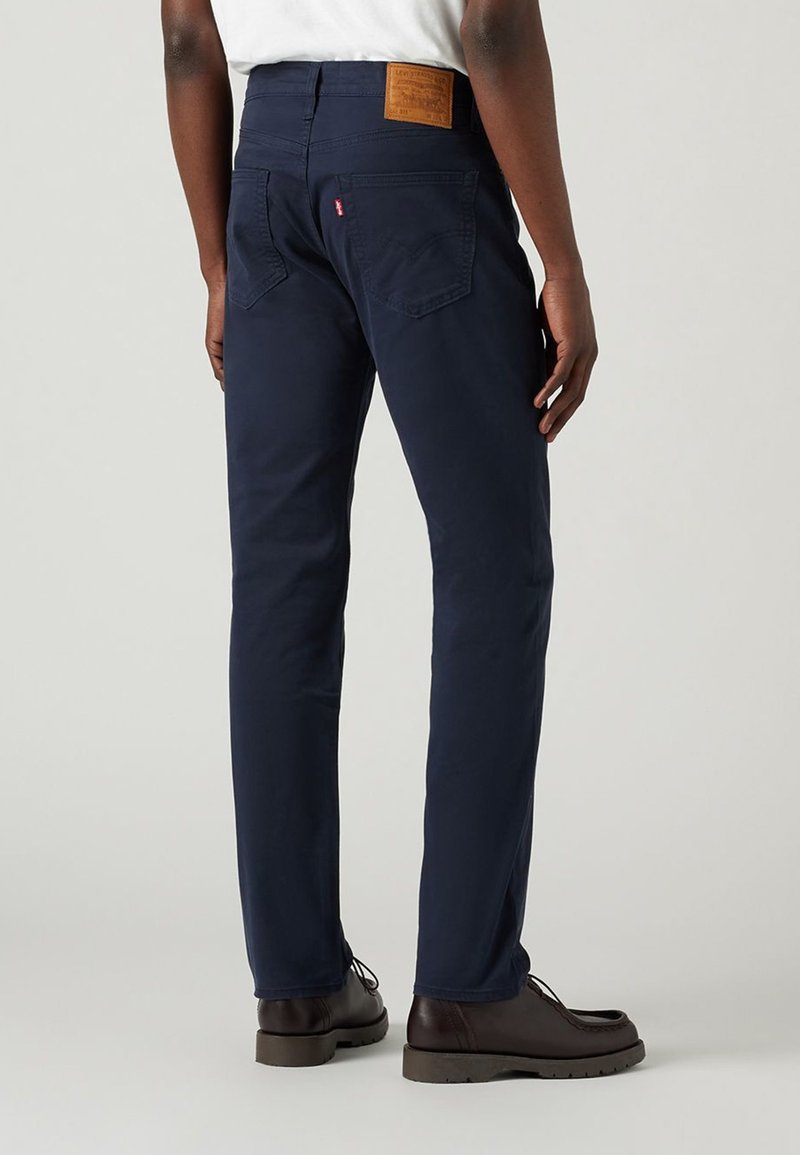 Pantalones chinos azul marino de algodón con corte recto. Presentan dos bolsillos traseros, un parche de cuero marrón en la cinturilla y zapatos marrones.