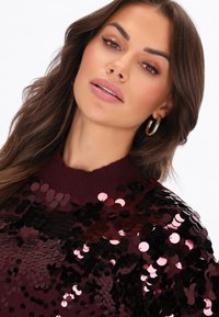 Donna con capelli castano ondulati indossa un maglione bordeaux decorato con grandi paillettes scure e orecchini a cerchio dorati, guardando leggermente verso l'alto.