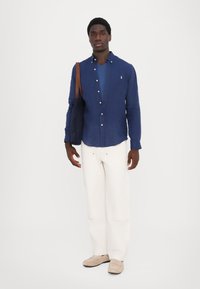 Model v obleki z modro srajco s gumbi čez temno modro majico z V-izrezom, belimi širokimi hlačami in bež loaferji, drži temno torbo.