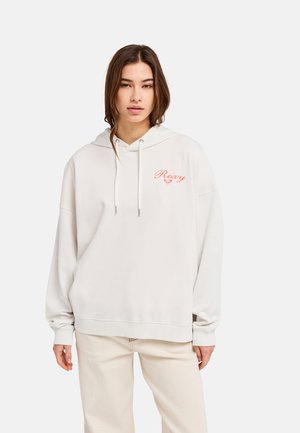 Junge Frau trägt ein weißes, übergroßes Hoodie mit dem orangefarbenen "Roxy"-Logo auf der Brust und hellbeige Hosen vor einem einfarbigen Hintergrund.