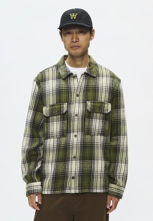 Homme portant une chemise à carreaux verte et blanche avec des poches à rabat et une casquette noire avec un logo jaune sur un fond uni.