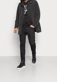 Giacca nera impermeabile con cappuccio e numerose tasche, abbinata a una felpa nera, jeans neri e sneakers nere con suola bianca.