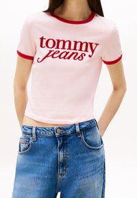 Pink cropped t-shirt with red contrast collar and sleeves, featuring “tommy jeans” in bold red text. Paired with high-waisted blue denim jeans.